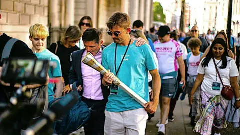 A man holding the Baton of  Hope during an event in Kent in 2023 with another man putting his arm around him and others in the background