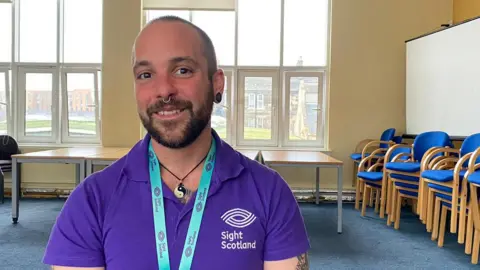 A man in his 30s wearing a purple t-shirt smiling at the camera. He has very short hair, a beard and a moustache. 