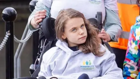 Karen Ryan Emily Ryan is seen wearing a grey hoodie and a black T-shirt.
Emily is pictured using her wheelchair and her mother, Karen, is seen standing behind her.
