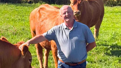 Lee Caine, wearing a grey T-shirt, standing next to his Limousin cattle in a field.