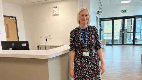 Carly West-Burnham the hospital's director of strategy stands in front of the new reception desk inside the Community Diagnostic Centre. She is wearing a black dress decorated with red cherries and white blossom. She has a blonde bob and is wearing and NHS ID card around her neck. 