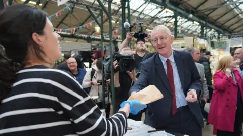 PA Media Hilary Benn who has grey hair and glasses and is wearing a suit is given a paper bag containing food by a woman stall holder with black hair at St George's Market 