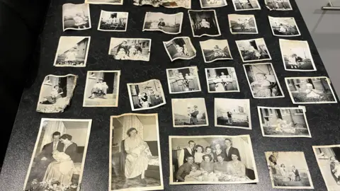 William Lindsay Family photos are spread out across a kitchen table, showing black and white images of children and families as well as a few colour images.