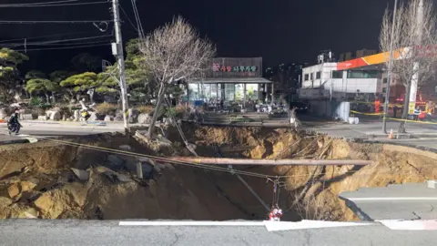 NEWS1 A massive sinkhole in the middle of a road in Seoul at night, in front of a convenience store and a petrol station. An exposed underground pipe stretches across the width of the hole and a tree on the edge of the sinkhole looks like it's about to fall in.