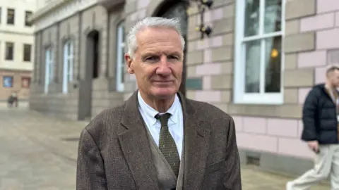 A man in a suit outside the States of Jersey building. The building has pink walls.