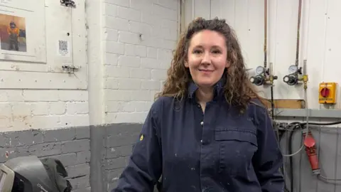 Nicola Ellis, who is wearing navy overalls covered in flecks of plaster, is smiling at the camera. There is a table to the right of her with a protective helmet on it. 