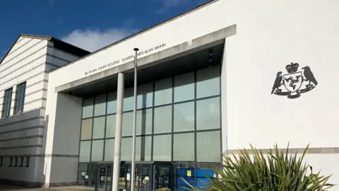 The outside of the court building, which is large, white and glass-fronted. the government crest is on the right.