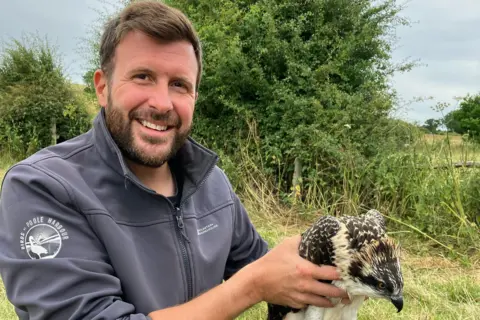 Birds of Poole Harbour Paul Morton smiles towards the camera as he holds an osprey ready for release. He has short brown hair and a full short beard. He is wearing a grey zip-up jacket with a white round Birds of Poole Harbour logo on the shoulder.