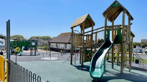East Lindsey District Council A play area filled with play equipment. On the right-hand side there is a wooden structure with steps and ladders, a bridge from one side to another, and a green slide. On the left-hand side, a roundabout and another slide.