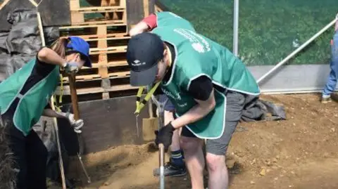 St Andrew's Healthcare A man and a woman both wearing dark green St Andrew's Healthcare jackets and caps, dig holes with spades in the earth. 