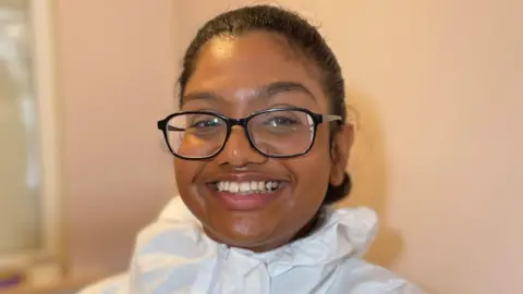 Daveena is wearing a white overall and smiling