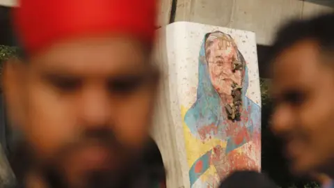 NurPhoto via Getty Images A painting of Sheikh Hasina wearing a blue scarf and with Bangladesh flags beneath her on a concrete column appears to have holes in it, and is covered in splats of red paint. The blurry face of a man in a red hat can be seen in the foreground