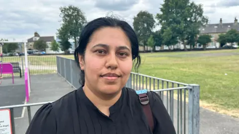 Malcolm Ring/BBC A woman with tied-back black hair. She is wearing a black top, and is standing in front of the play area of a park. In the distance is grass and a number of trees.