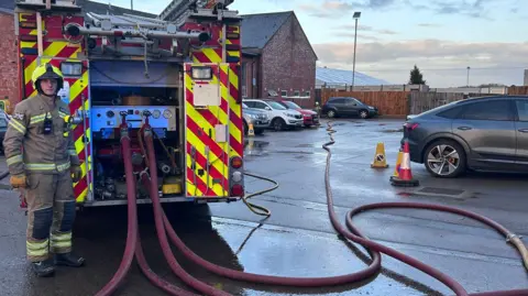 Oxfordshire Fire and Rescue Service A firefighter stands at the rear of one of the fire engines which has four hoses leading off towards the building.
