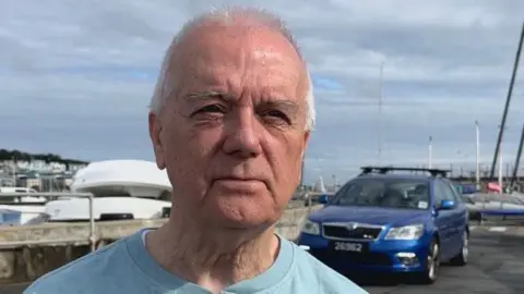 Aindre Reece Sheerin is standing outside. Boats and parked cars are behind him. He is wearing a blue shirt. 