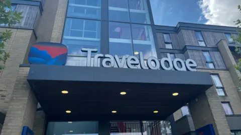 Emma Baugh/BBC A Travelodge hotel on Newmarket Road in Cambridge. There is a Travelodge sign and logo on the entrance of the building.