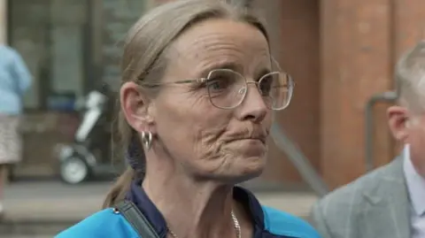 Joanne Moody standing outside a redbrick building, wearing large specs on her face, hoop earrings, a necklace, and a blue sports jersey. The background is softly blurred and includes steps leading to the building entrance and a partially visible mobility scooter.