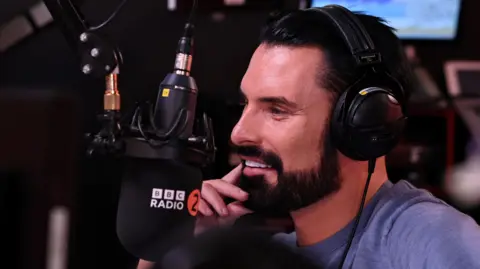 Rylan - who has a black beard and hair and white teeth. He's wearing a blue T-shirt and has a Radio 2 microphone in front of him.