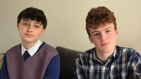 Paul Moseley/BBC Arlo is on the left side of the image wearing blue sweater over a white-collared shirt. George, on the left, has a blue and white checked shirt. They are sitting on a sofa.