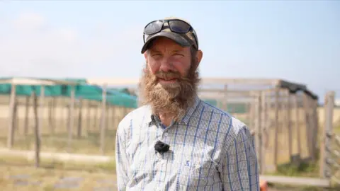Simon Higgs posa para la cámara al aire libre. Detrás de él se ve un recinto para pájaros en un campo.