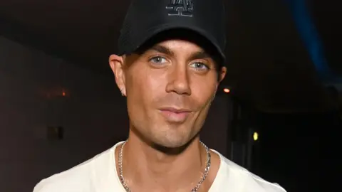 Max George looking into the camera while wearing a baseball cap