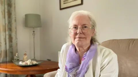 A woman with shoulder-length grey hair wearing a white cardigan and lilac scarf sits in her living room. She is on a light brown armchair and behind her is a brown coffee table with shells on a plate and two statues next to it on the table. There is a lamp with a light-green shade in the corner of the room, next to light gold and green floral patterned curtains. 