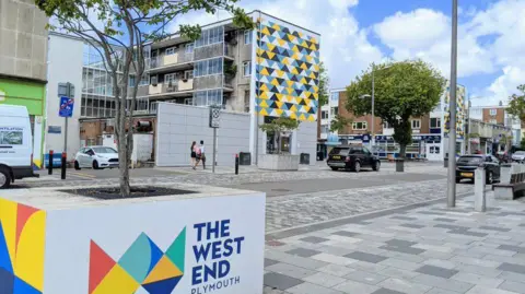 Plymouth's West End area. It contains a number of buildings which are up to four or five storeys high. There are several trees and parked cars along the road.