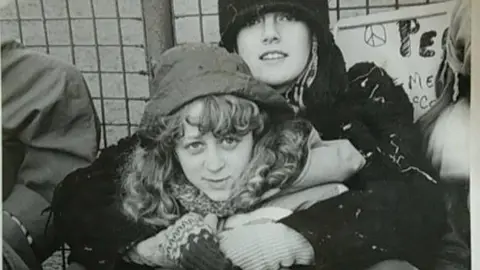 Emma Woolf A black and white photo of two women sitting on the ground with their arms around each other. One woman is wearing a black hat. The other woman has curly hair and is also wearing a hat. There are protest posters on a fence behind them, with peace symbols on them.