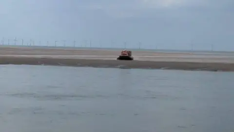 HM Coastguard A yellow RNLI hovercraft can be seen in the distance on the Leasowe sands with water in the foreground and wind turbines in the background.
