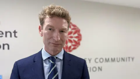 BBC Jim Roberts looking at the camera. Jim has short curly brown hair and is wearing a suit with a diagonally striped tie.