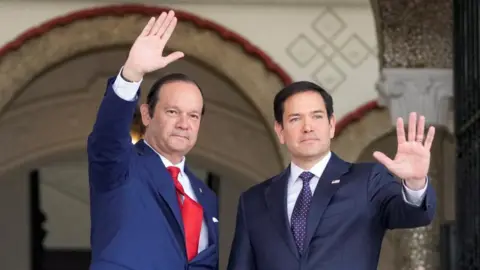 Reuters US Secretary of State Marco Rubio waves with Panamanian foreign minister Javier Martinez-Acha after arriving at the presidential palace in Panama City. Both men are wearing blue suits.