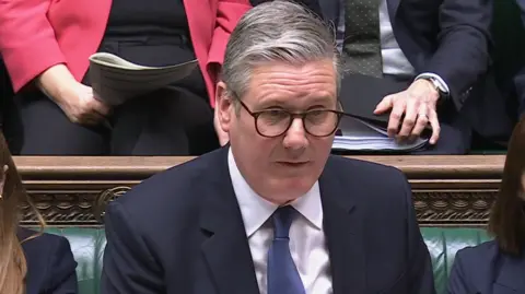 UK Parliament Sir Keir Starmer, wearing dark-rimmed glasses and a dark blue suit and blue tie, talking in the House of Commons.