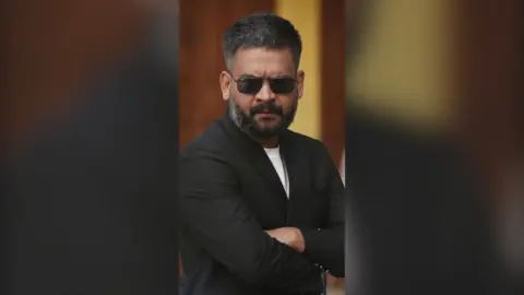 Man wearing black shades and a black blazer folds his arms.