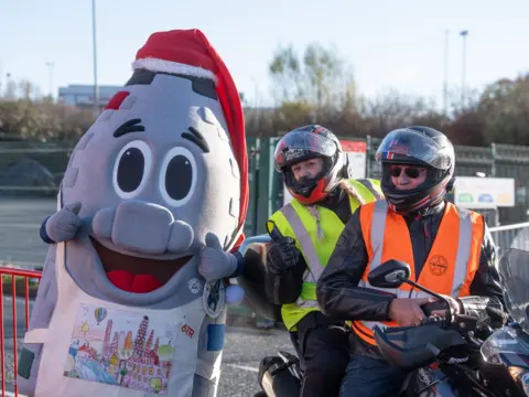 Ian Knight / Z70 Photography Stoke-on-Trent's centenary mascot, Kelvin the Kiln, appears next to two people in high visibility clothing sat on a bike. The mascot represents a traditional bottle kiln used in the ceramics industry, and is wearing a red Santa hat.