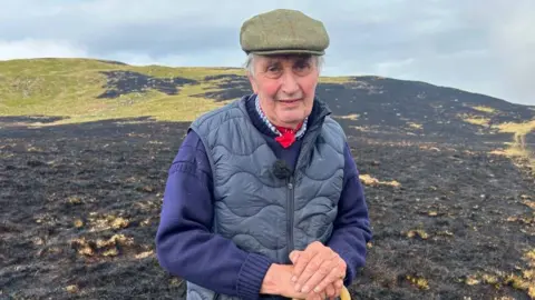 Brynmor Morgan who is wearing a blue jacket and green hat, standing in front of a heavily scorched field