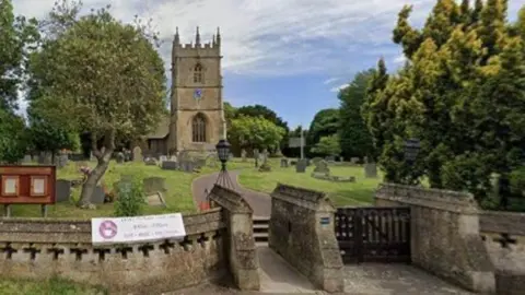 Google A village church and graveyard, with a stone path leading to the entrance and headstones scattered across the grass under a cloudy sky
