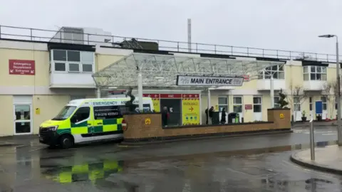 The main entrance to Frimley Park hospital which has a glass canopy over the doors and is a two storey building rendered in a pale yellow.