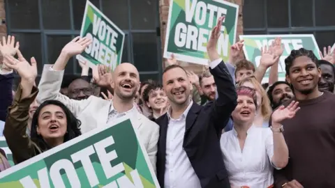 PA A mixed group of smartly dressed people wave with their hands in the air. Many in the crowd hold "vote green" signs. 