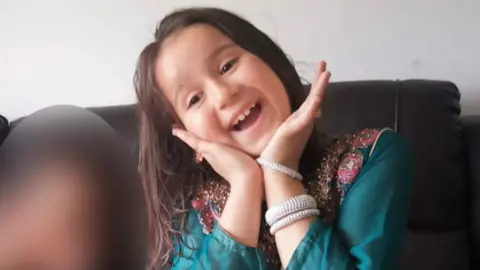 Sara Sharif smiling and looking directly at the camera. She is wearing a blue outfit and white bracelets. 