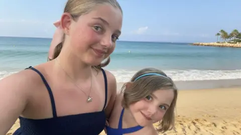 A selfie shows Ellie (left) on a beach with her younger sister Emilia (right). They are wearing summer dresses on a sunny day and the sand and sea are behind them.