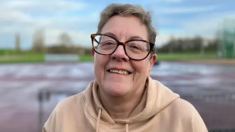 A woman with short grey hair and glasses, wearing a light brown hoodie, smiles at the camera.