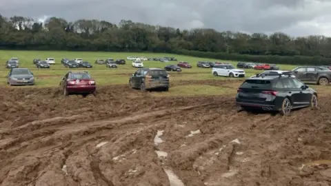 PA Media A very muddy field on which several cars are parked. Some of the cars are splattered with mud. There are deep tyre marks in the mud.