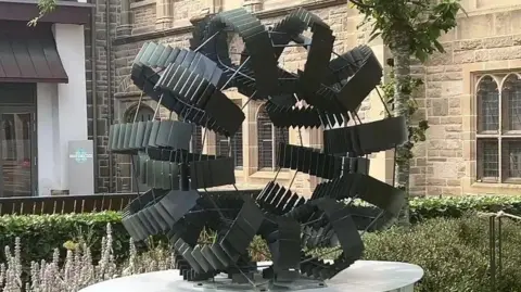 Circular sculpture featuring tyres made of black slate-like items in a Blackburn public garden. A hedge is surrounding the sculpture and there's an old stone-built building in the background.