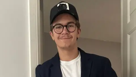 A young man, Freddie Penrose, is wearing a cap and glasses leans against a door frame. He is wearing a suit jacket.