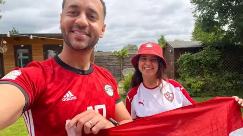A couple dressed in Egyption football artier, holding a flag and taking a selfie, with big smiles. They are in a garden. 