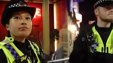 Two uniformed police officers, one male and one female, stand in front of the neon signs of a Manchester nightspot