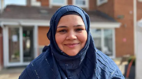 Shaista Khan is wearing a blue headscarf and is stood behind the mosque building, which is visible in the background.