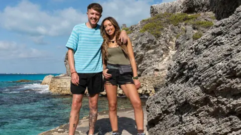 StudioLambert/BBC Roman Kemp has him arm around sister Harleymoon as they both smile on a path next to the sea next to a large rock.
