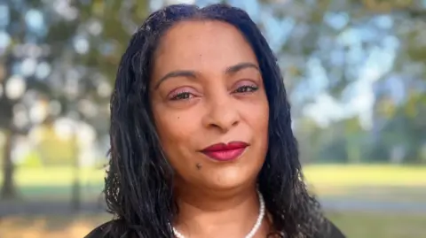 Alda Simoes looking into the camera. She has long dark hair and is wearing a white pearl-style necklace and is standing outside on a sunny day