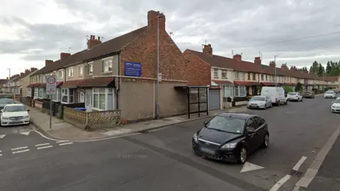 Longford Street in Middlesbrough. There is a bus stop fixed on to side of a house and there is a black car travelling in one direction. There are several rows of terraced houses. 